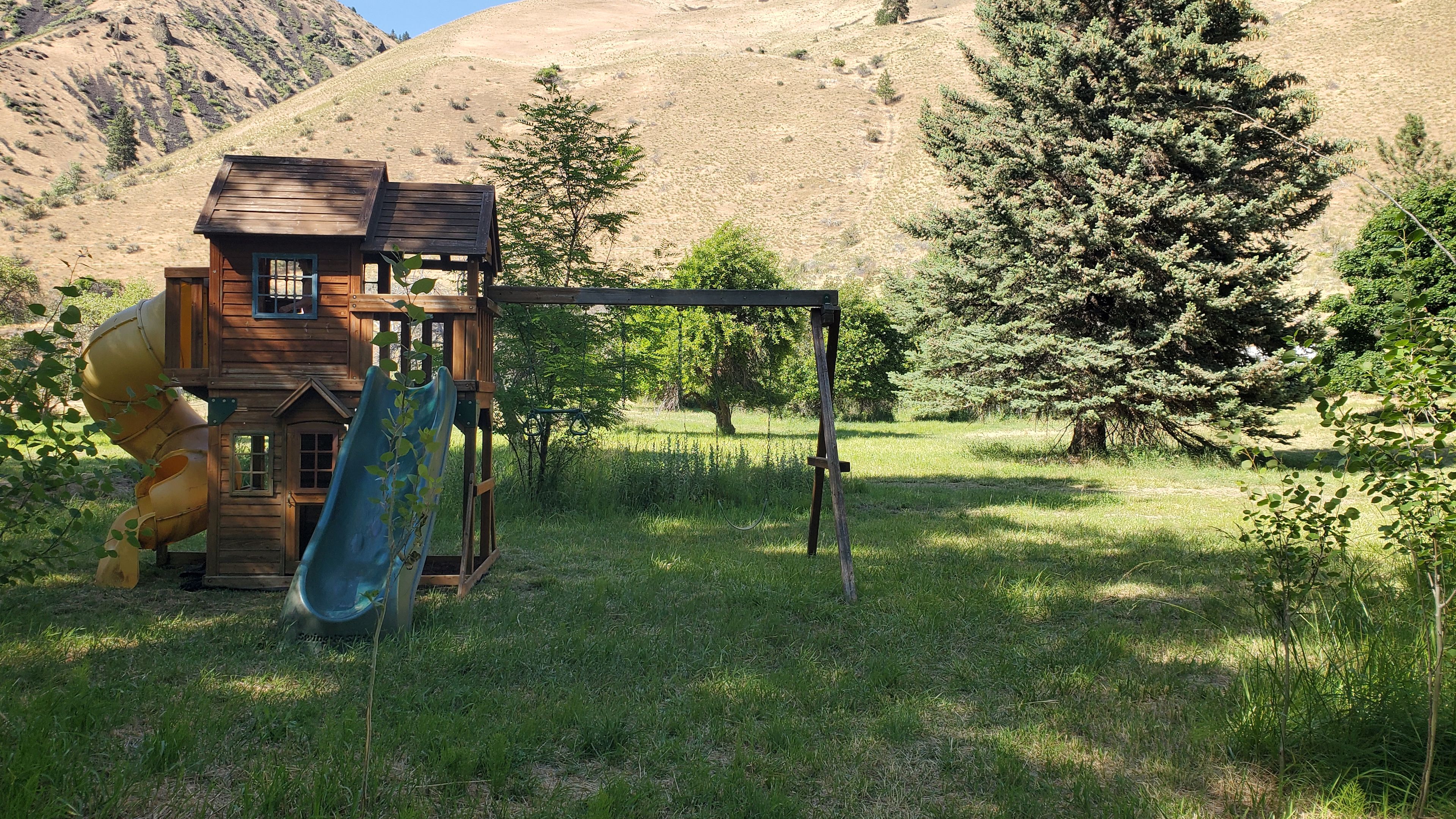 Une structure de jeu en bois, dotée d’un toboggan et d’une zone d’escalade, entourée d’herbe et d’arbres.
