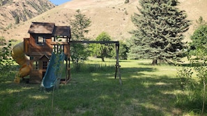 Une structure de jeu en bois, dotée d’un toboggan et d’une zone d’escalade, entourée d’herbe et d’arbres.