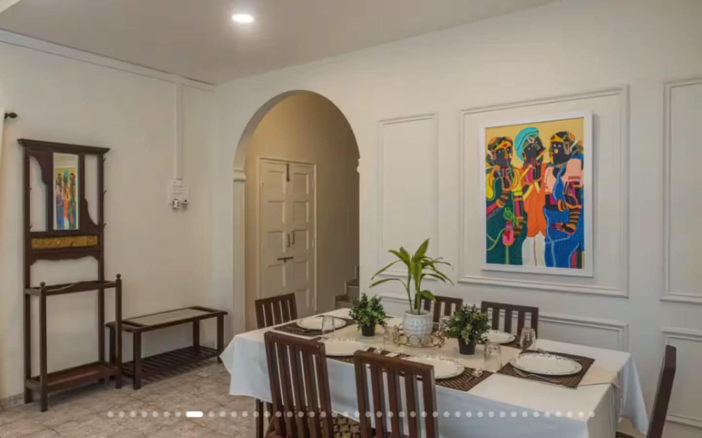 A dining room with a white table set for six, surrounded by chairs. There is a painting on the wall, a door leading to another room, and a small table with a plant.