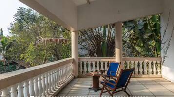 A covered balcony with a white balustrade, a table with chairs, and lush greenery in the background.
