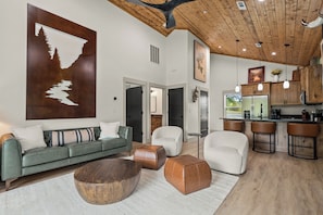 A modern living room with a large wooden coffee table, a sofa, and a white armchair. There is a kitchen area with bar stools and a refrigerator. - Bungalow Bliss | NEW | Swim Spa 💦 Private Fire Pit 🔥 (Hollister)