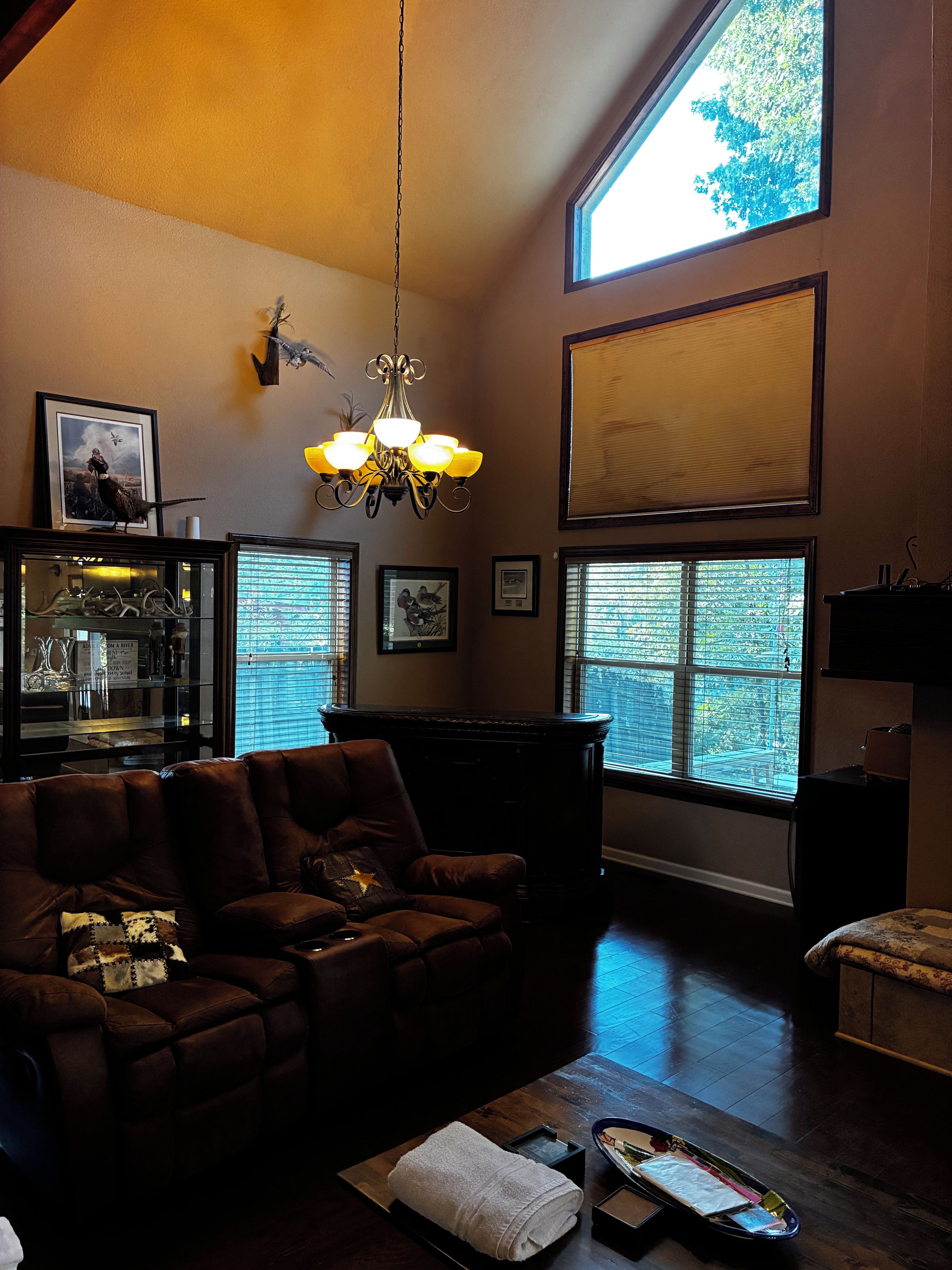 A living room with a brown leather sofa, a chandelier, and a large window with blinds.