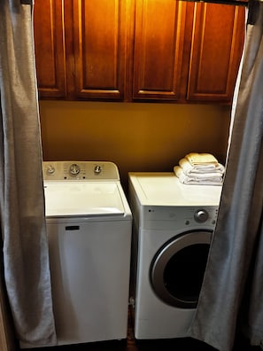 A laundry area with a front-loading washer and dryer, wooden cabinets above, and a curtain partially drawn.