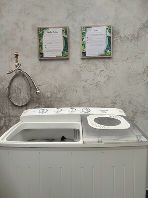 Máquina de lavar de abertura frontal com painel de controle, uma mangueira e dois cartazes informativos na parede.