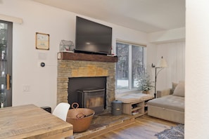 Ein gemütliches Wohnzimmer mit Kamin aus Stein, einem darüber angebrachten Flachbildfernseher, einem Holzesstisch und einer bequemen Couch.