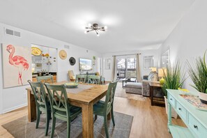 A spacious living area with a dining table, chairs, a sofa, and a coffee table. There is a large window providing natural light and a view of the outdoors. - Seabrook Exclusives | 2013 Marsh Walk (Seabrook Island)