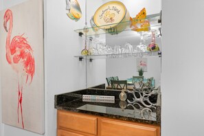 A kitchen with a granite countertop, a glass shelf with decorative items, and a painting of a flamingo. - Seabrook Exclusives | 2013 Marsh Walk (Seabrook Island)