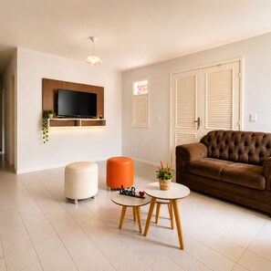 A modern living room with a flat-screen TV mounted on the wall, a brown sofa, an orange ottoman, and two round tables with a small plant on one. - Holiday home with capacity for up to 10 people, 30m from the shopping center. (Piranhas)