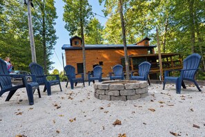 A cabin with a stone fire pit surrounded by Adirondack chairs, set in a forested area.