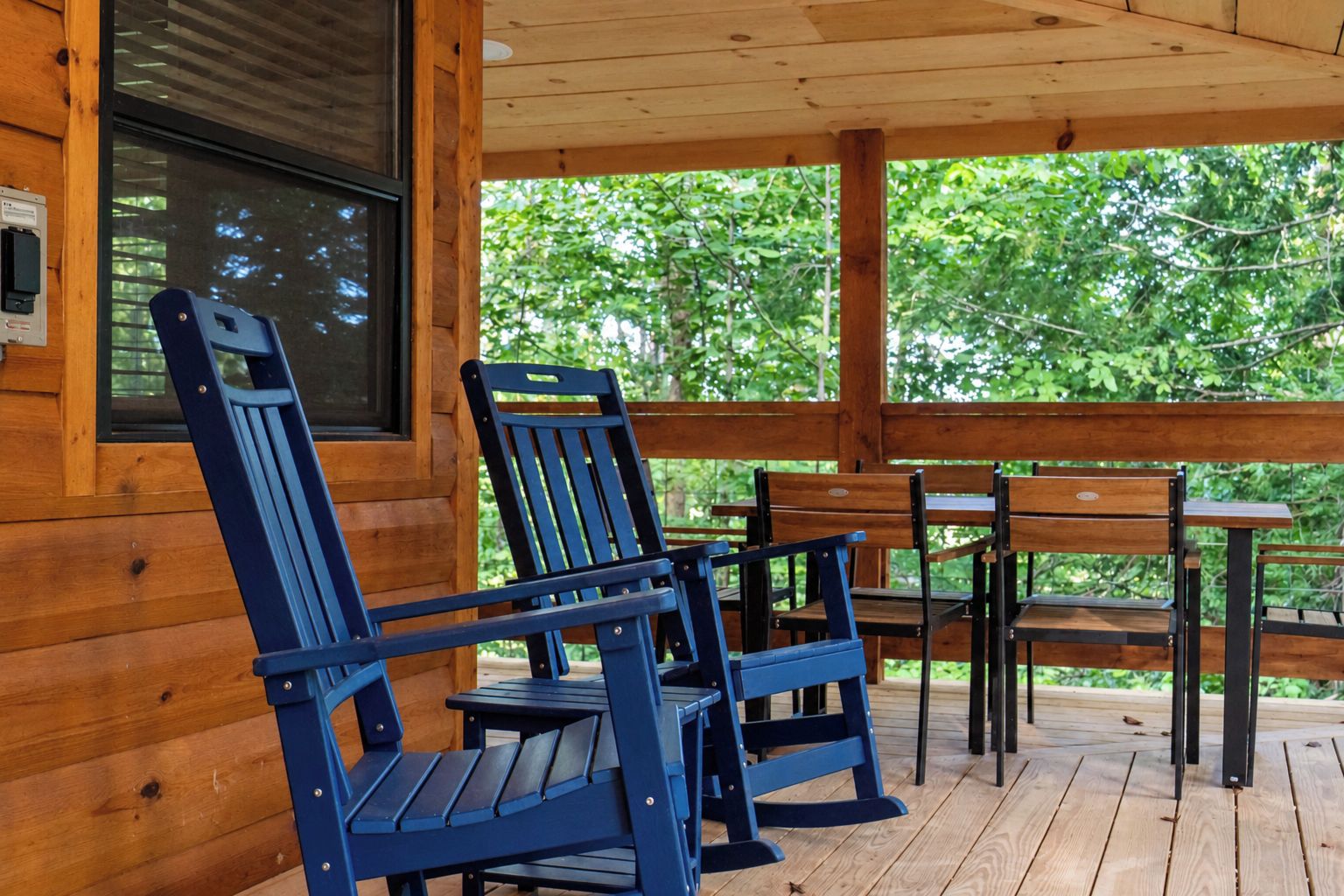 A wooden deck with a table and chairs, a window, and a view of greenery.