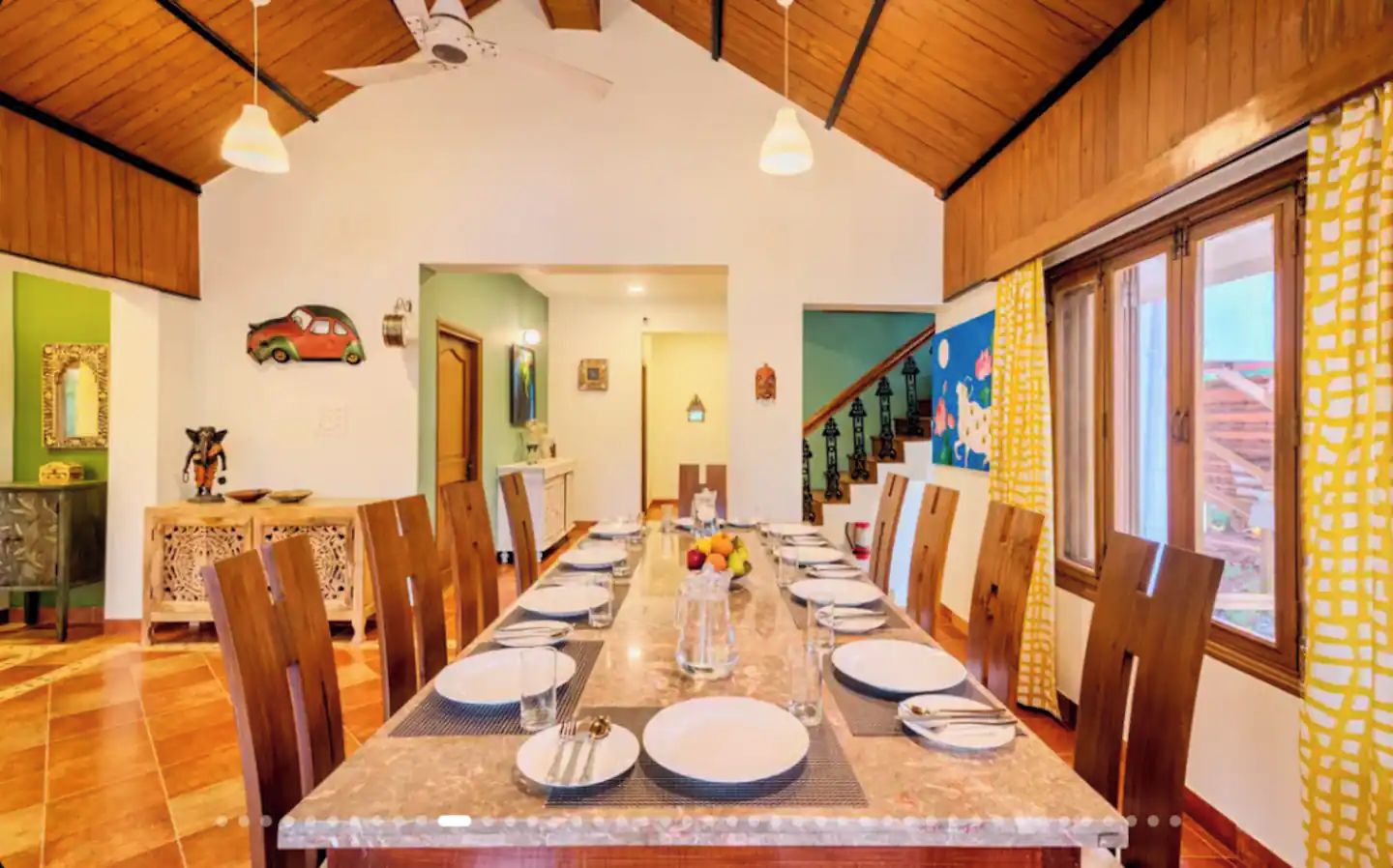 A dining area with a long table set for multiple people, wooden chairs, and a view of the outside through large windows.