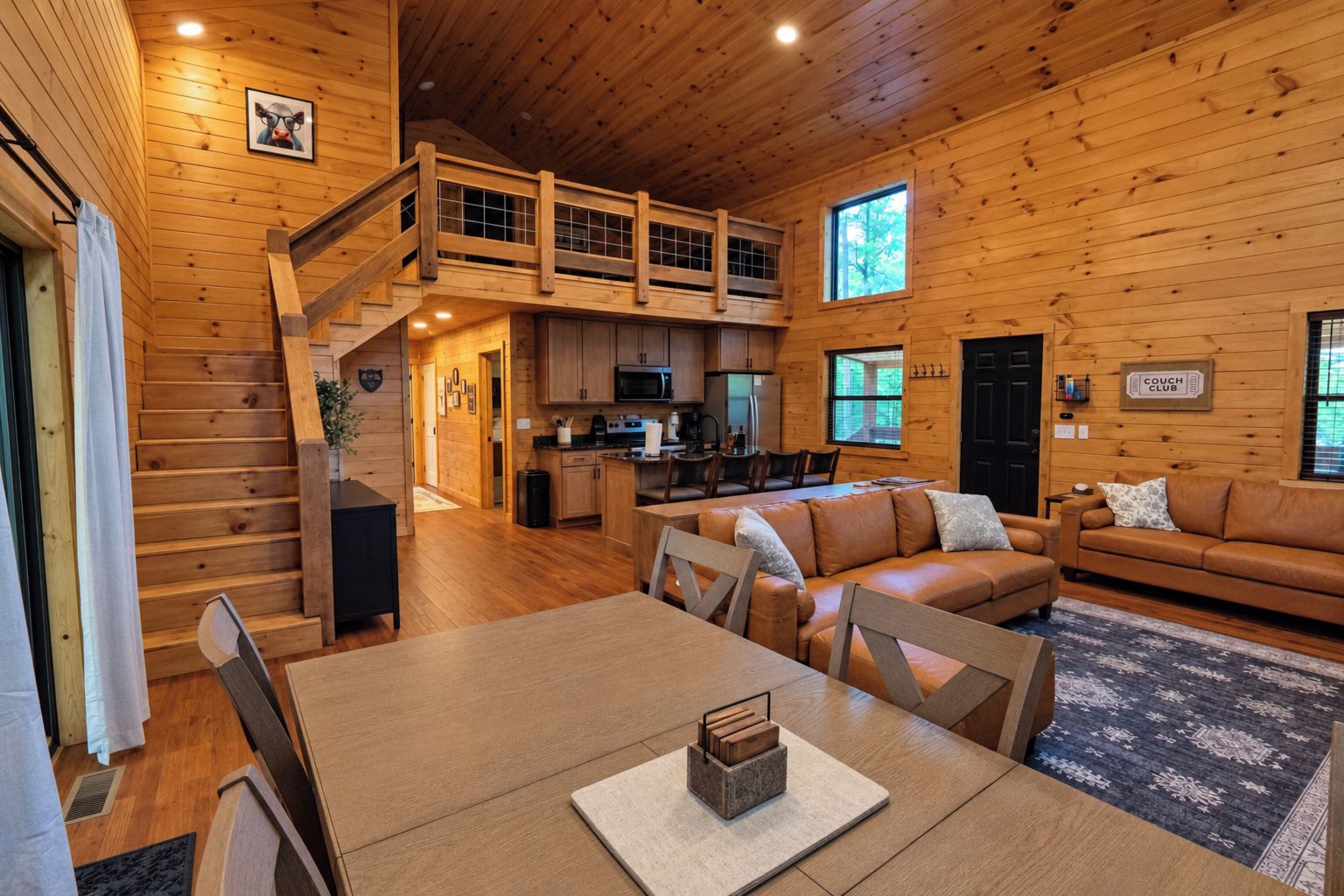 A cabin interior with a dining area, a living room with a sofa, a kitchen, and a staircase leading to an upper level.