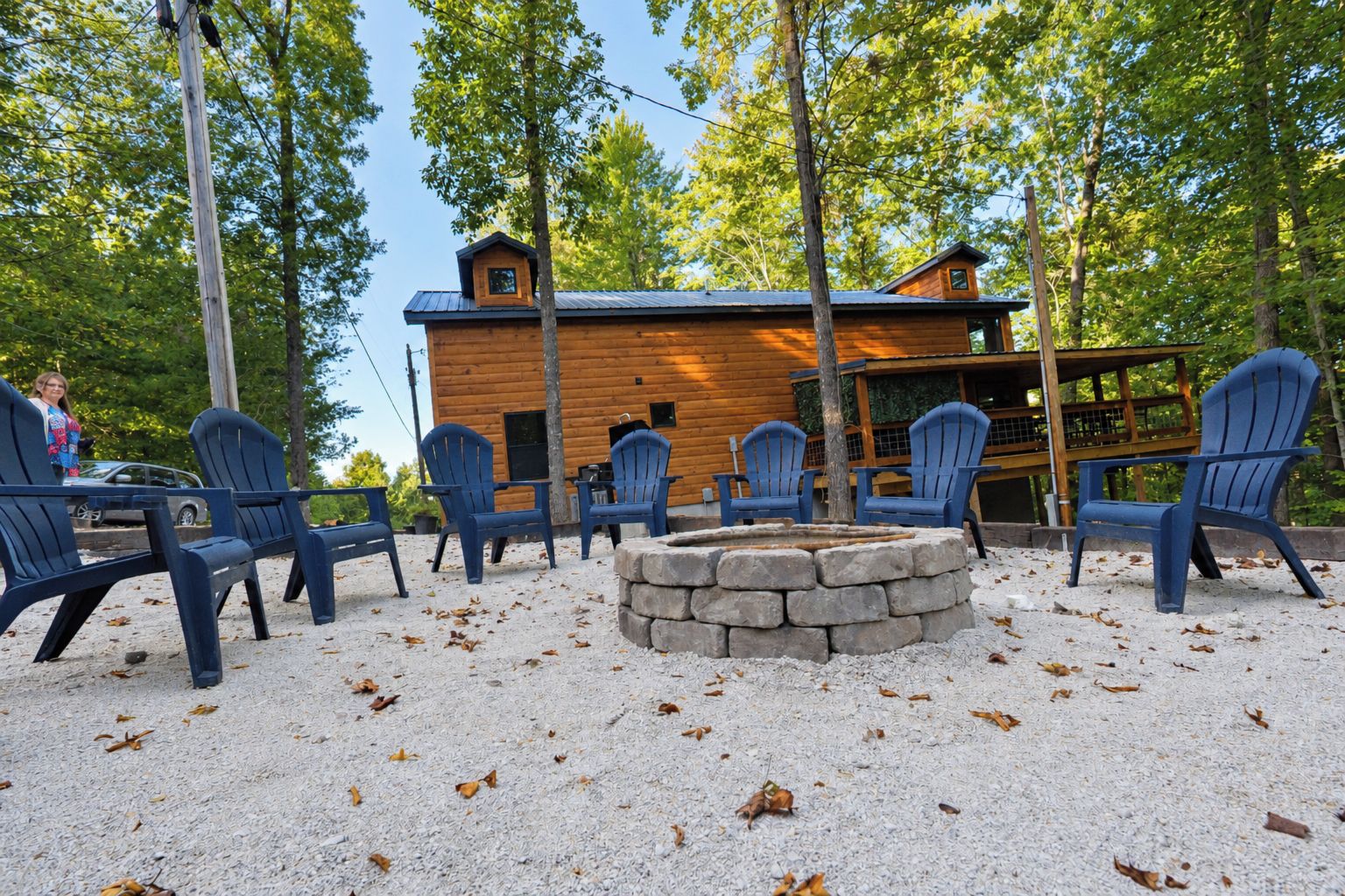 A cabin with a stone fire pit surrounded by Adirondack chairs, set in a forested area.