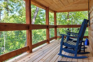 A wooden deck with a rocking chair and a table, surrounded by a wooden railing, overlooking a forest.