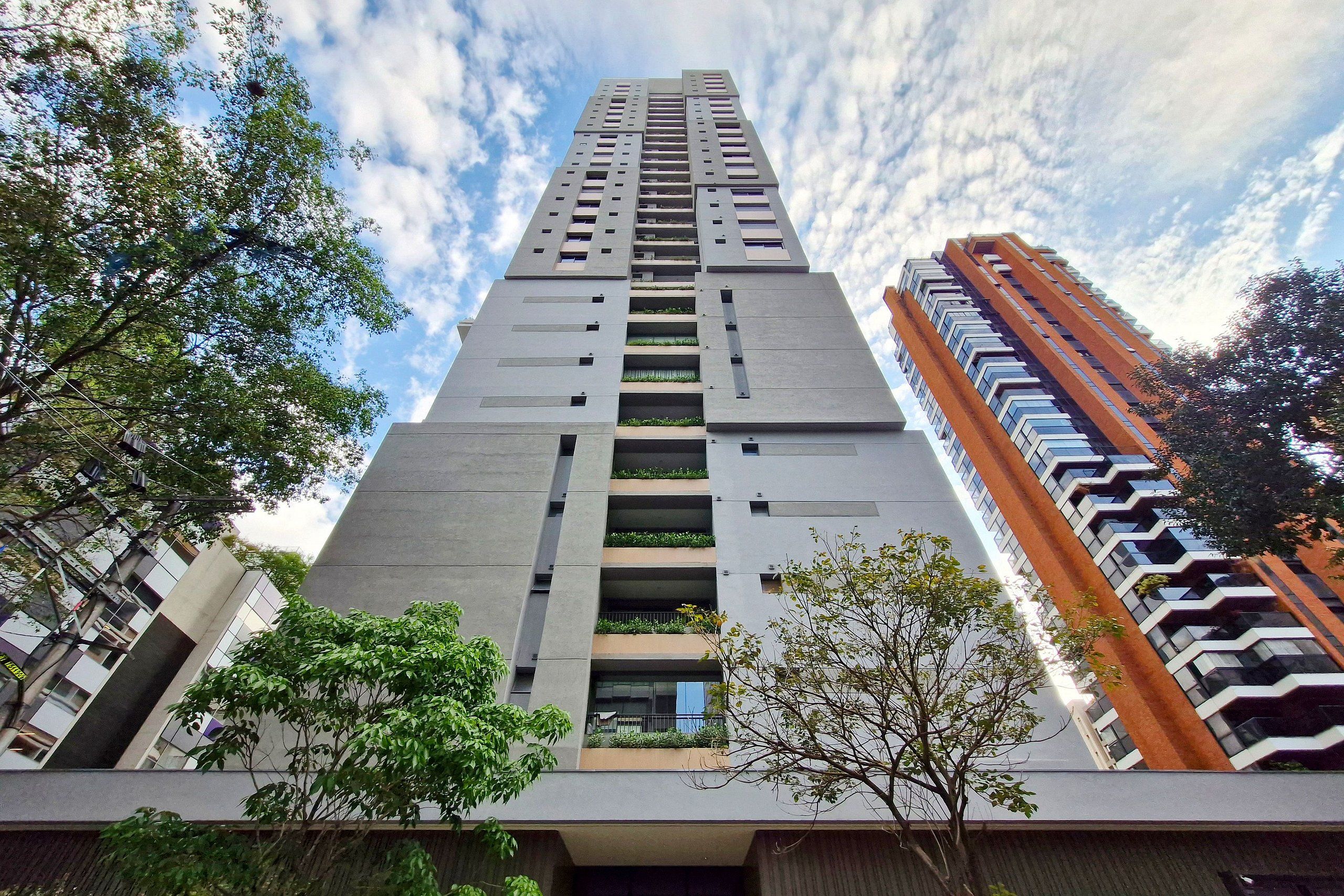 A tall, modern building with a grey facade, multiple balconies, and a mix of greenery.