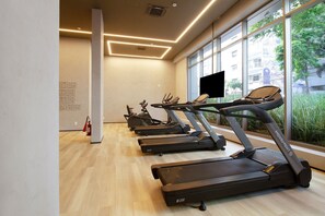 Ein Fitnessstudio mit mehreren Laufbändern, einem Feuerlöscher und einem großen Fenster mit Blick auf Gebäude und Grünflächen.