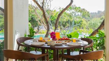 A dining table with plates, cutlery, and a glass of orange juice, set against a backdrop of lush greenery and a pool.