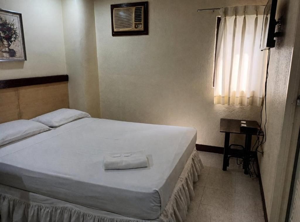 Une chambre d’hôtel avec un lit, une petite table, une chaise, un climatiseur fixé au mur et une fenêtre avec des rideaux.