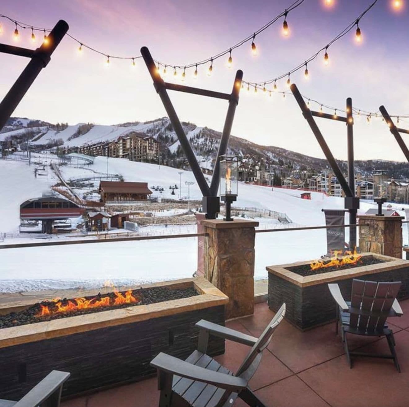 An outdoor seating area with fire pits and string lights, overlooking a snowy landscape with buildings and a ski slope.