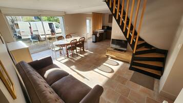 A living room with a brown sofa, a dining table with chairs, a TV on a wooden stand, and a staircase with wooden steps.