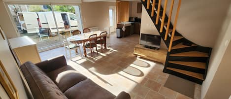 A living room with a brown sofa, a dining table with chairs, a TV on a wooden stand, and a staircase with wooden steps.