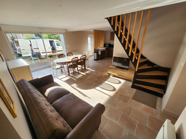 A living room with a brown sofa, a dining table with chairs, a TV on a wooden stand, and a staircase with wooden steps.
