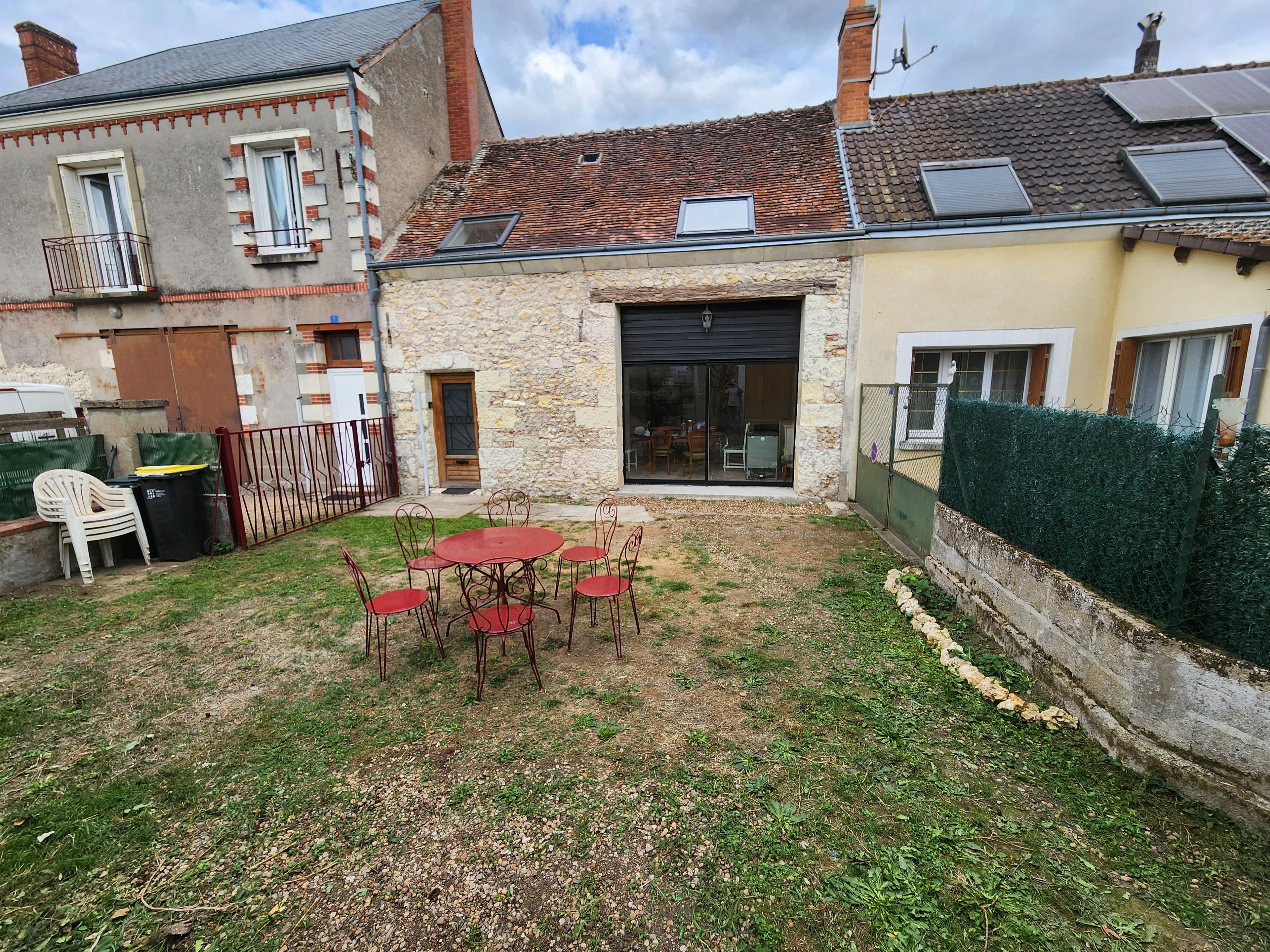 Une cour avec une table et des chaises rouges, un bâtiment en pierre et un espace délimité par une clôture.