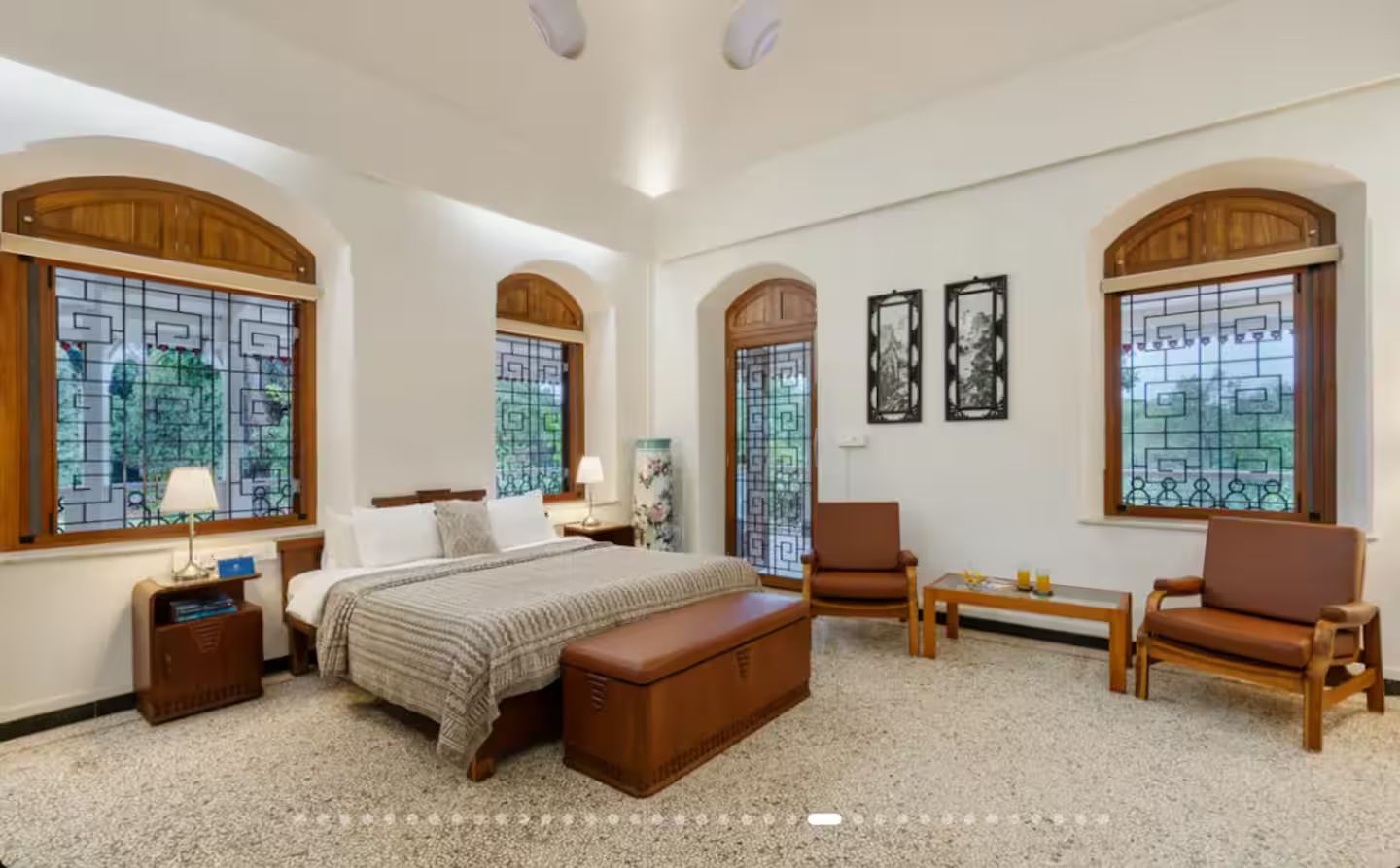 Ein Hotelzimmer mit einem großen Bett, zwei Sesseln, einem kleinen Tisch und einer Bank. Es gibt gewölbte Fenster mit dekorativen Glasverzierungen und gerahmte Kunstwerke an den Wänden.