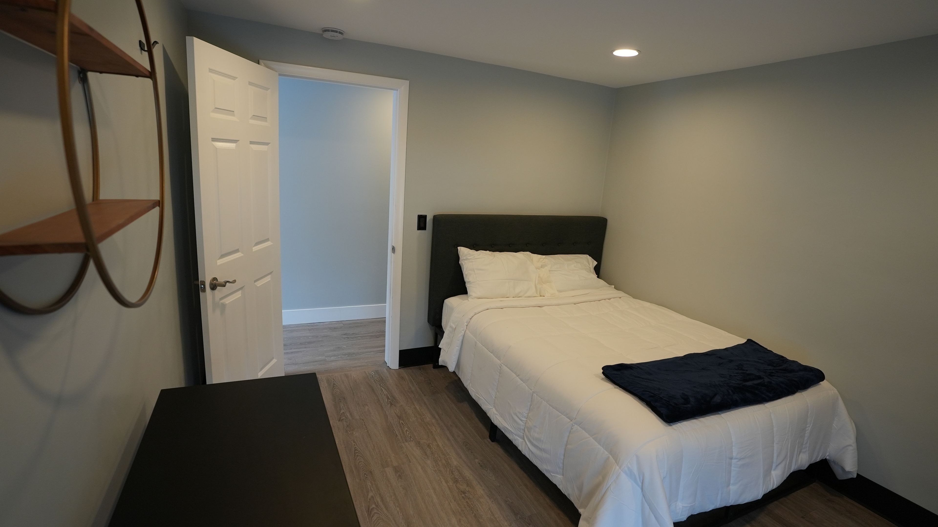 A bedroom with a bed, a door, a bench, and a wall-mounted shelf.