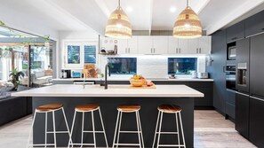 A modern kitchen with a central island, bar stools, and a large window. - Riverbank Blue - luxe pool, sauna, cabana and family fun moments near Minnamurra's best beaches (Minnamurra)