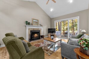 A living room with a fireplace, a television, and a large window overlooking a forest. - Woodhaven - Deer Park 39 (Glen Arbor)