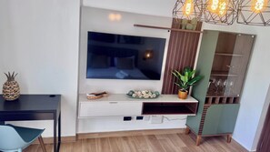 A modern living room with a flat-screen TV mounted on a wall, a wooden coffee table, a chair, a small plant, and a decorative shelf with glasses. - Earthing Central Park Suiteconnect and Rest (Punta Cana)