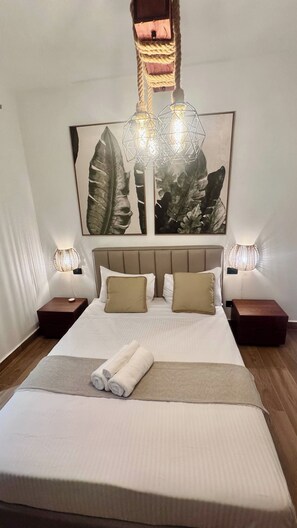 Una cama bien hecha con cabecera, dos mesitas de noche con lámparas y una obra de arte grande con motivos de hojas sobre la cama.