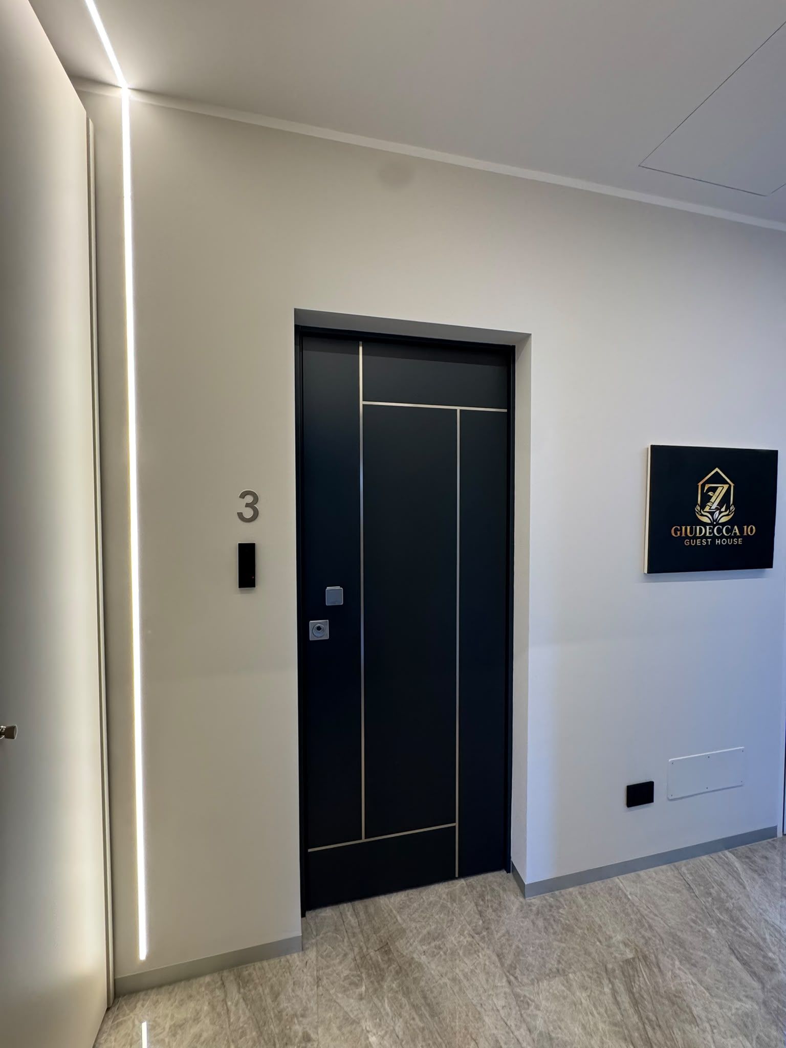 Un ascenseur moderne, avec une façade noire et un écran numérique, une enseigne murale portant le logo « GIUDECCA 10 GUEST HOUSE » et le chiffre « 3 » indiquant l’étage.