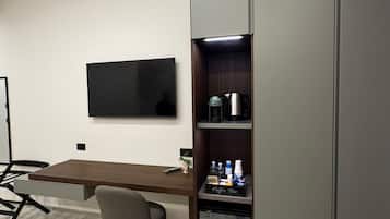 Un espace de bureau moderne doté d’un téléviseur à écran plat fixé au mur, d’un bureau en bois avec une chaise, et d’un coin café intégré comprenant une bouilloire et une machine à café.