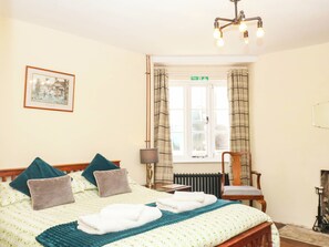 Ein Hotelzimmer mit einem Bett, einem Sessel, einer Lampe, einem fenster mit Vorhängen und einem gerahmten Bild an der Wand.