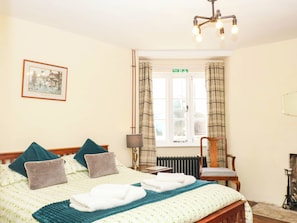 Ein Hotelzimmer mit einem Bett, einem Sessel, einer Lampe, einem fenster mit Vorhängen und einem gerahmten Bild an der Wand.