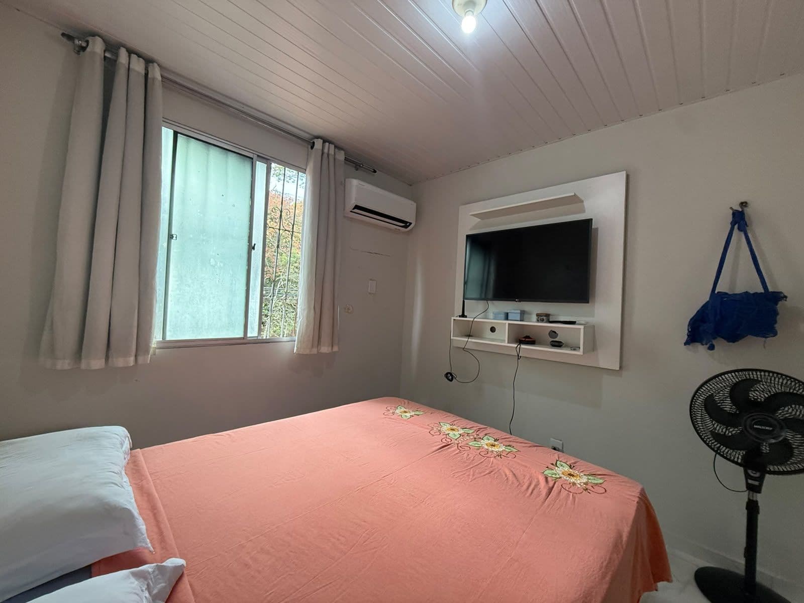 Un dormitorio con cama, ventana con cortinas, aire acondicionado, televisor montado en la pared, ventilador y una bolsa azul colgada en la pared.