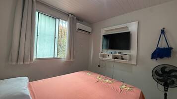 Un dormitorio con cama, ventana con cortinas, aire acondicionado, televisor montado en la pared, ventilador y una bolsa azul colgada en la pared.