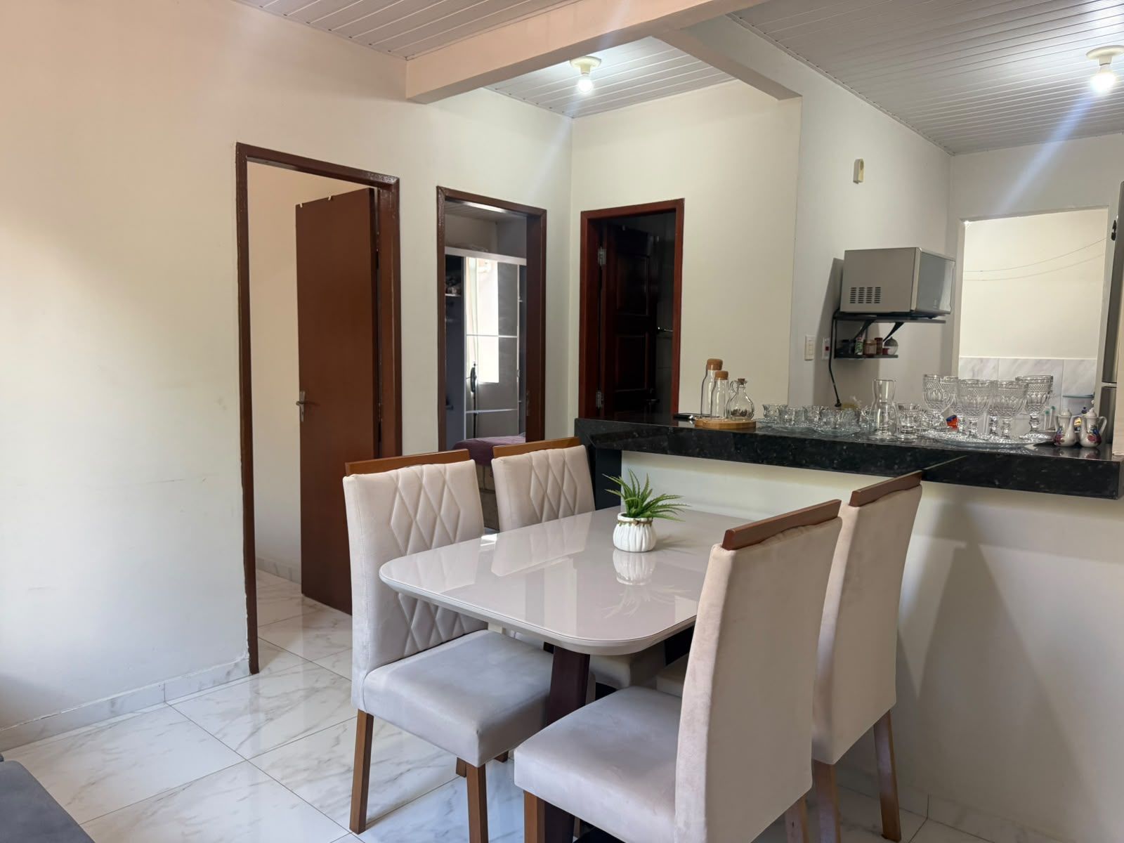 Un comedor con una mesa y sillas blancas, una planta en maceta y una cocina con armarios y microondas.