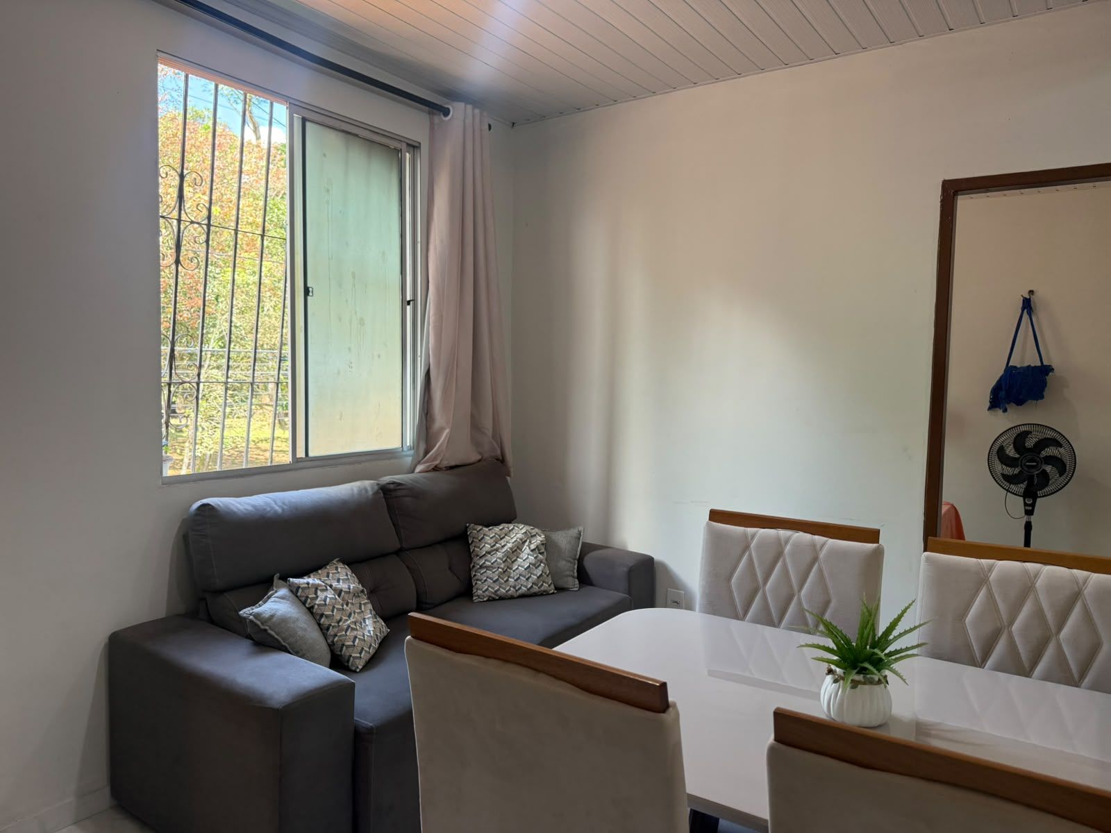 Una sala de estar moderna con un sofá, una mesa de comedor y sillas. Hay una ventana con cortina, un ventilador de pared y una planta pequeña sobre la mesa.