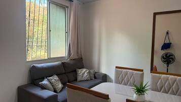 Una sala de estar moderna con un sofá, una mesa de comedor y sillas. Hay una ventana con cortina, un ventilador de pared y una planta pequeña sobre la mesa.