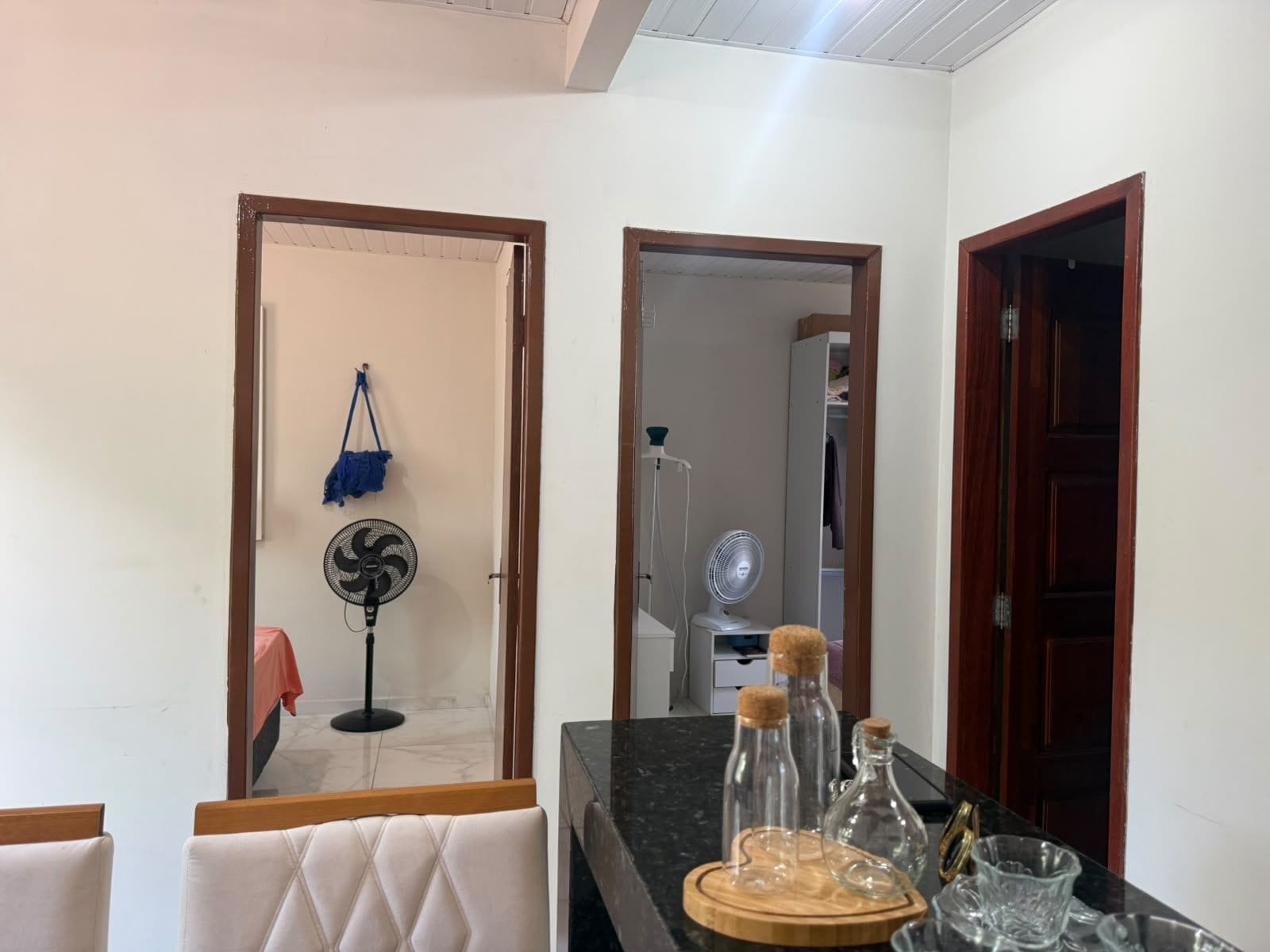 A room with a dining area, a kitchen counter with glass bottles, and a fan in the background.