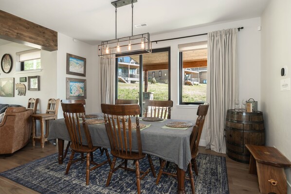 A dining area with a table set for four, wooden chairs, a blue patterned rug, and a view of a house through a glass door.