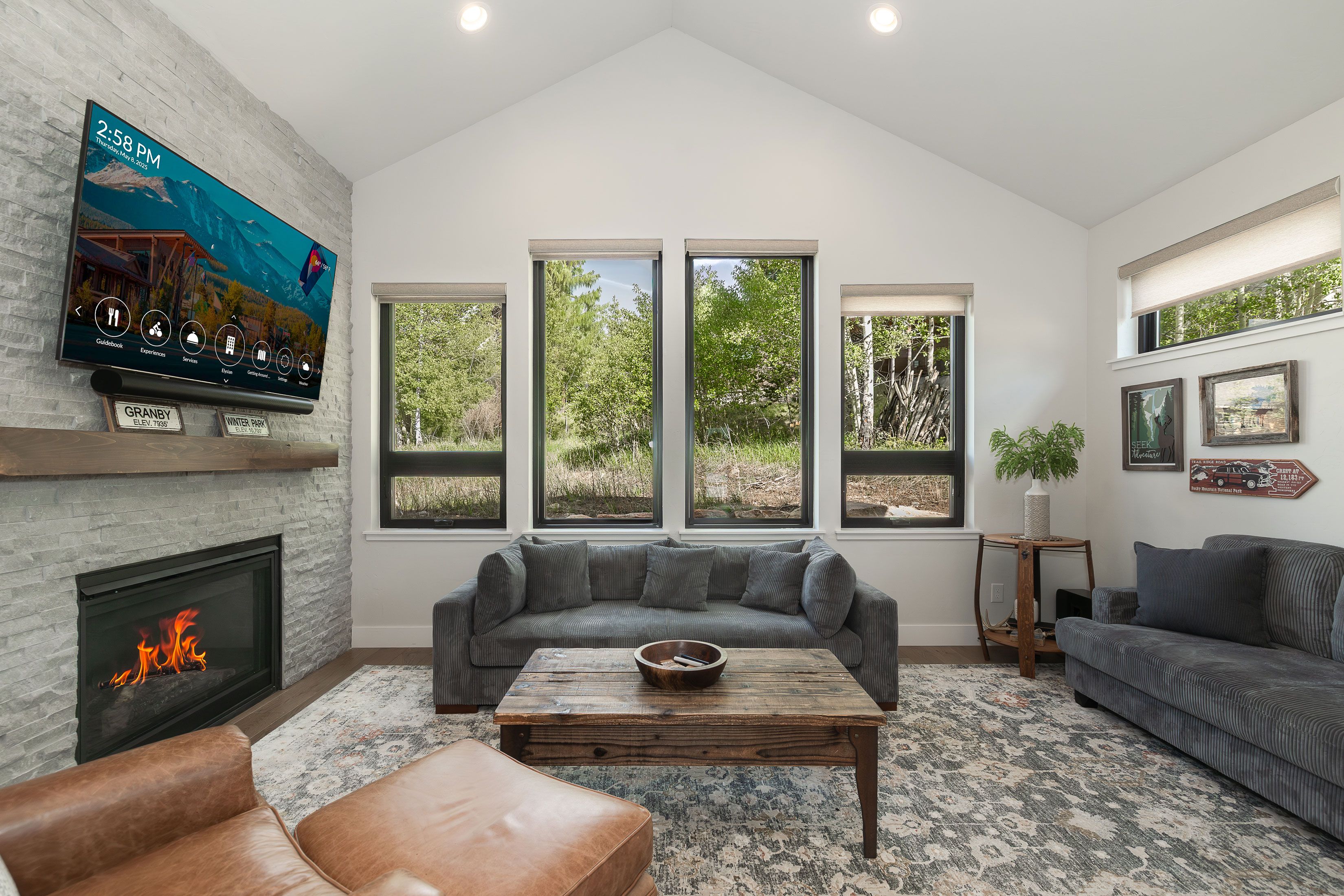 A modern living room with a fireplace, a large window, and a mounted television displaying a mountain scene.