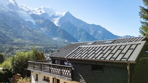Uma cabana na montanha com telhado inclinado, rodeada por montanhas arborizadas e um céu claro.