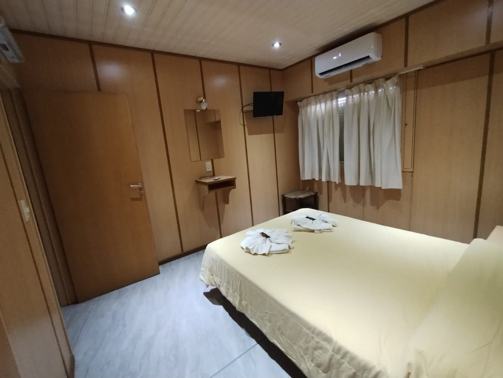 Een hotelkamer met een bed, een klein nachtkastje, een airconditioner, een spiegel en een raam met gordijnen.
