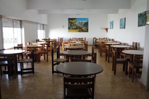 Un comedor amplio con varias mesas y sillas de madera, ventanas grandes con persianas y un cuadro en la pared.