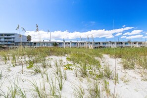 Un hôtel situé en bord de mer, composé de bâtiments à plusieurs étages, avec une plage de sable bordée de gazon et un ciel bleu dégagé.
