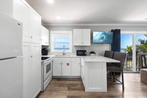 A modern kitchen with white cabinetry, a built-in oven, a microwave, and a sink. There's a dining area with a wicker chair and a table, and a large TV mounted on the wall displaying a beach scene. - Sunrise Ocean Views & Beach Access, 400 Ft to Downtown Cocoa Beach | 1BR Condo, Sleeps 4 (Cocoa Beach)